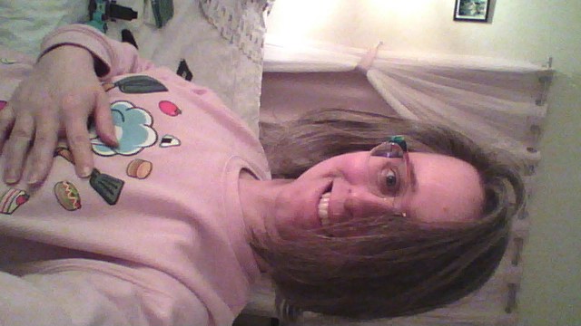 Selfie of me standing in my old bedroom wearing a pink Kirby sweater and smiling, my hair is almost covering half of my face.
