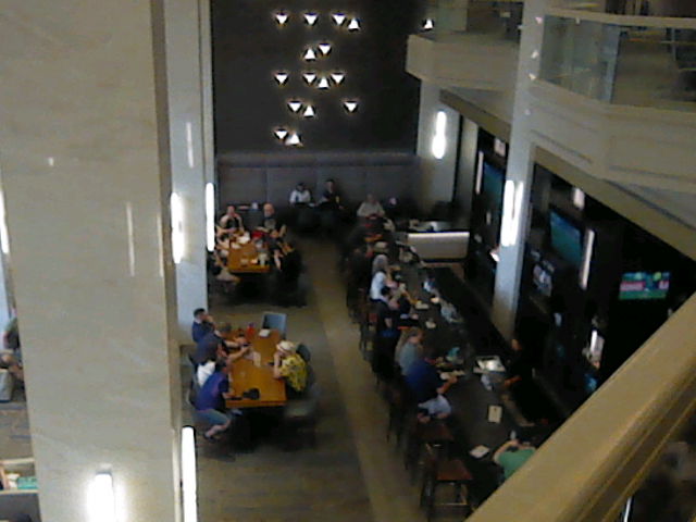 A shot of the lounge and bar at the SGDQ 2026 hotel in Minneapolis.  The photograph is taken from the mezzanine level and you can see a few more balcony-type out-croppings.  Below at the bar are people seated eating and drinking, and behind them are more people at tables.  They are framed on the left by some pillars.