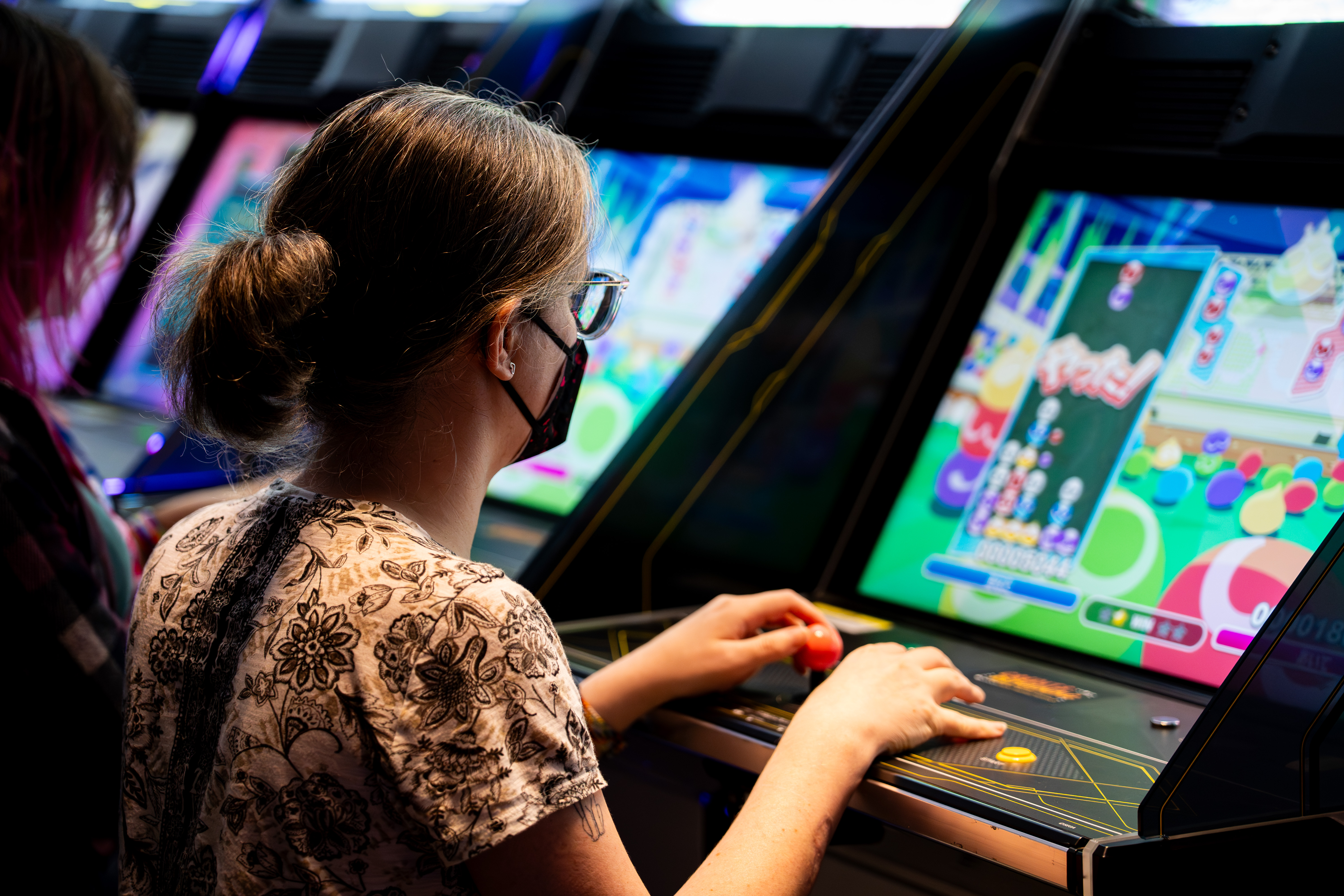 My wife and me, playing linked Puyo Puyo on arcade machines.  I am wearing a dark KN95 mask and my Puyo board is looking pretty healthy. My left hand delicately holds the joystick while my right is positioned over the buttons. 