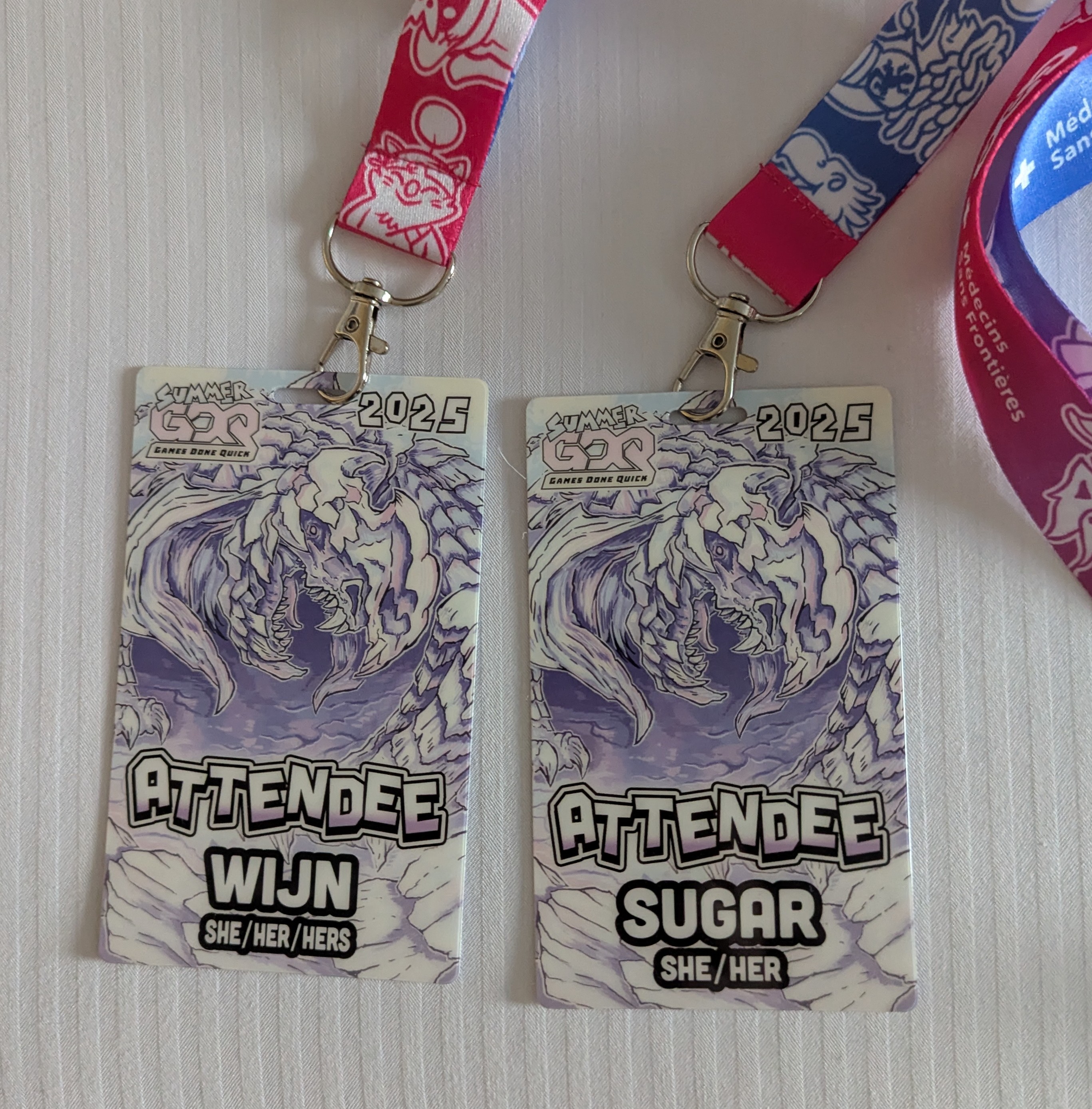 Two Attendee badges for Summer Games Done Quick. They have this pastel purple and white dragon art on them. They both say Attendee. My wife's has her handle "wijn" and pronouns "she/her/hers" and mine has my handle "sugar" and pronouns "she/her".  