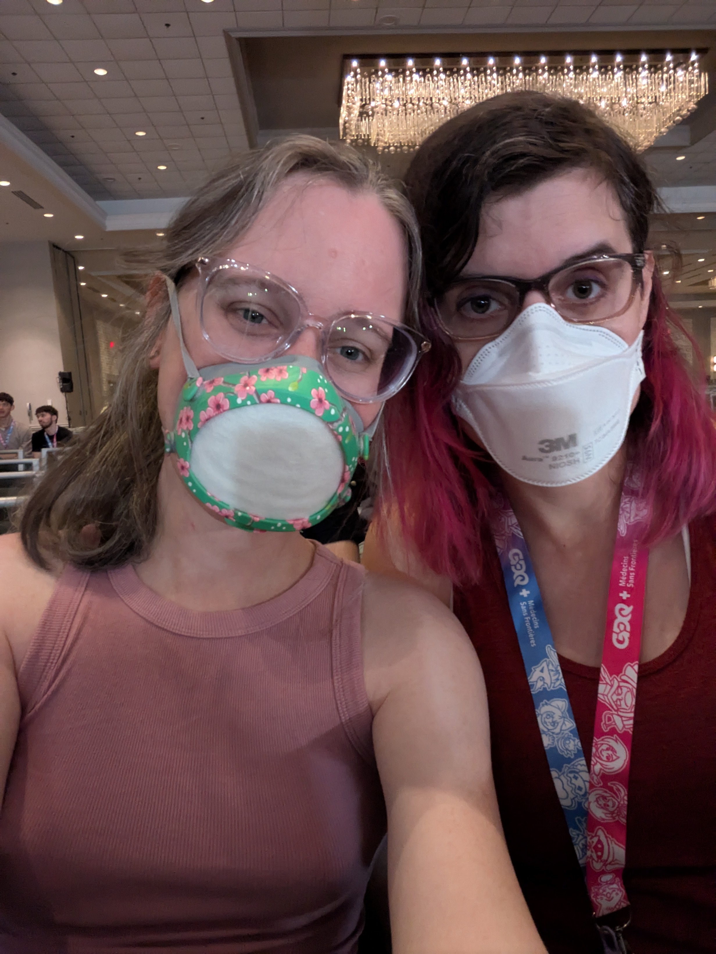 A selfie photo of my wife and me. We're both wearing masks, glasses, and tank tops. We're smiling with our eyes. We're in a big ballroom. 