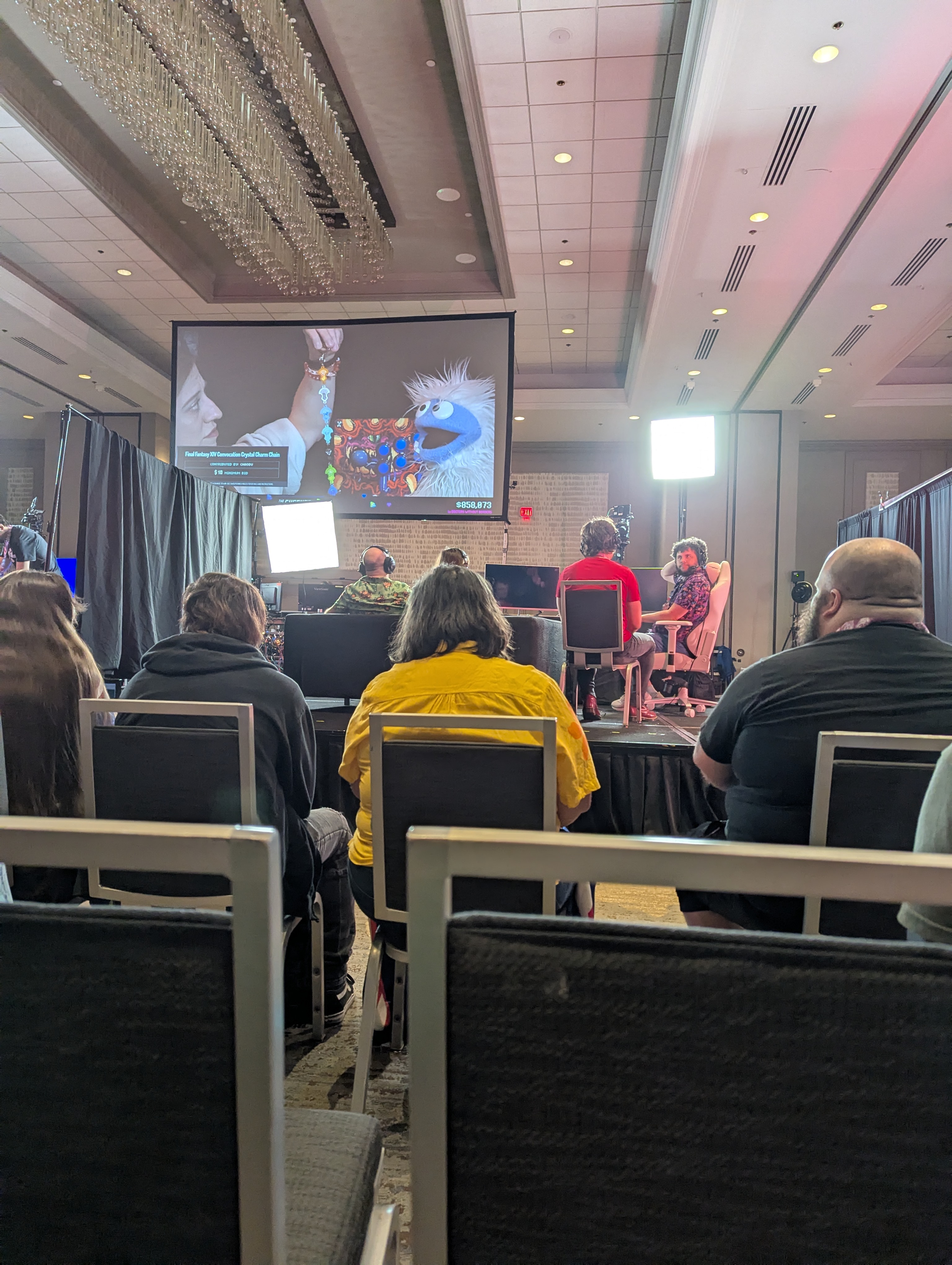 Photograph of my point of view at the time, looking to the stage where they are setting up for a new game run and on the projected screen behind them is a prize segment feature the mascot of The Yetee (merch partner), the Yetee, a blue and white abominable snowman muppet.