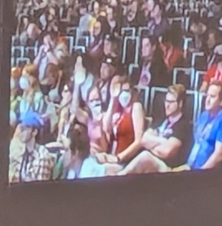 A photo of a projected screen showing a bunch of people sitting in chairs watching something else. Two of the women in the center are facing the camera and thus appear to be looking directly at the viewer. They're waving. It's us