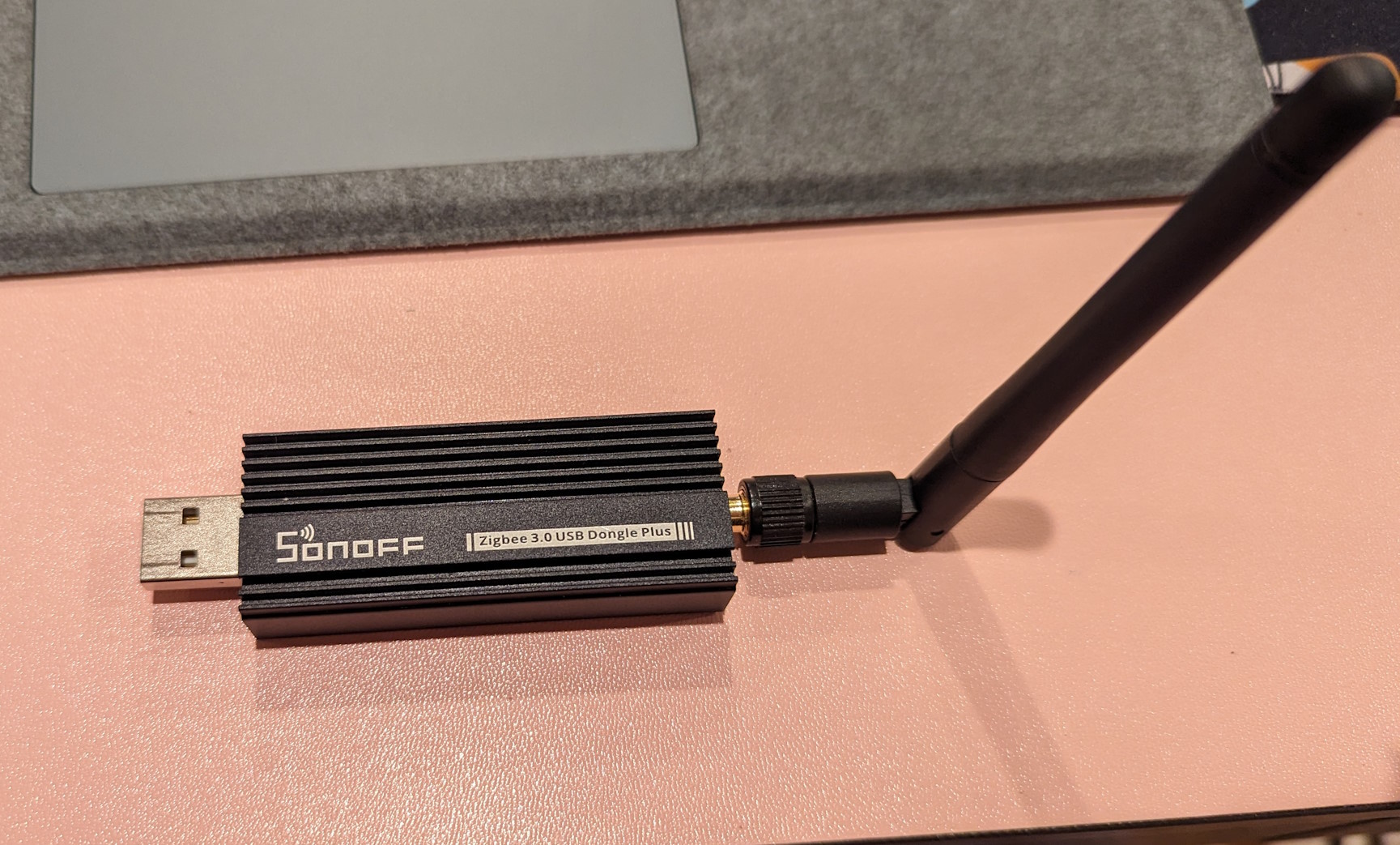 The SONOFF Zigbee 3.0 USB Dongle Plus sitting on a desk.  It is a USB device with a black metal chassis that also looks and acts as a heatsink.  On the left side is a USB port, on the right is an antenna.