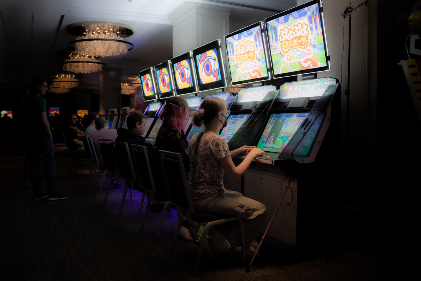A dark vignette shot of a row of sit down arcade machines, angled so that the machines are disappearing into the distance. sugar sits at the closest machine and is playing Puyo Puyo. To her left is her wife wijn playing the same game of Puyo Puyo on the second machine. They're playing against each other.  - Photo credit: Matt "Lowex" Lowe (official GDQ photographer)