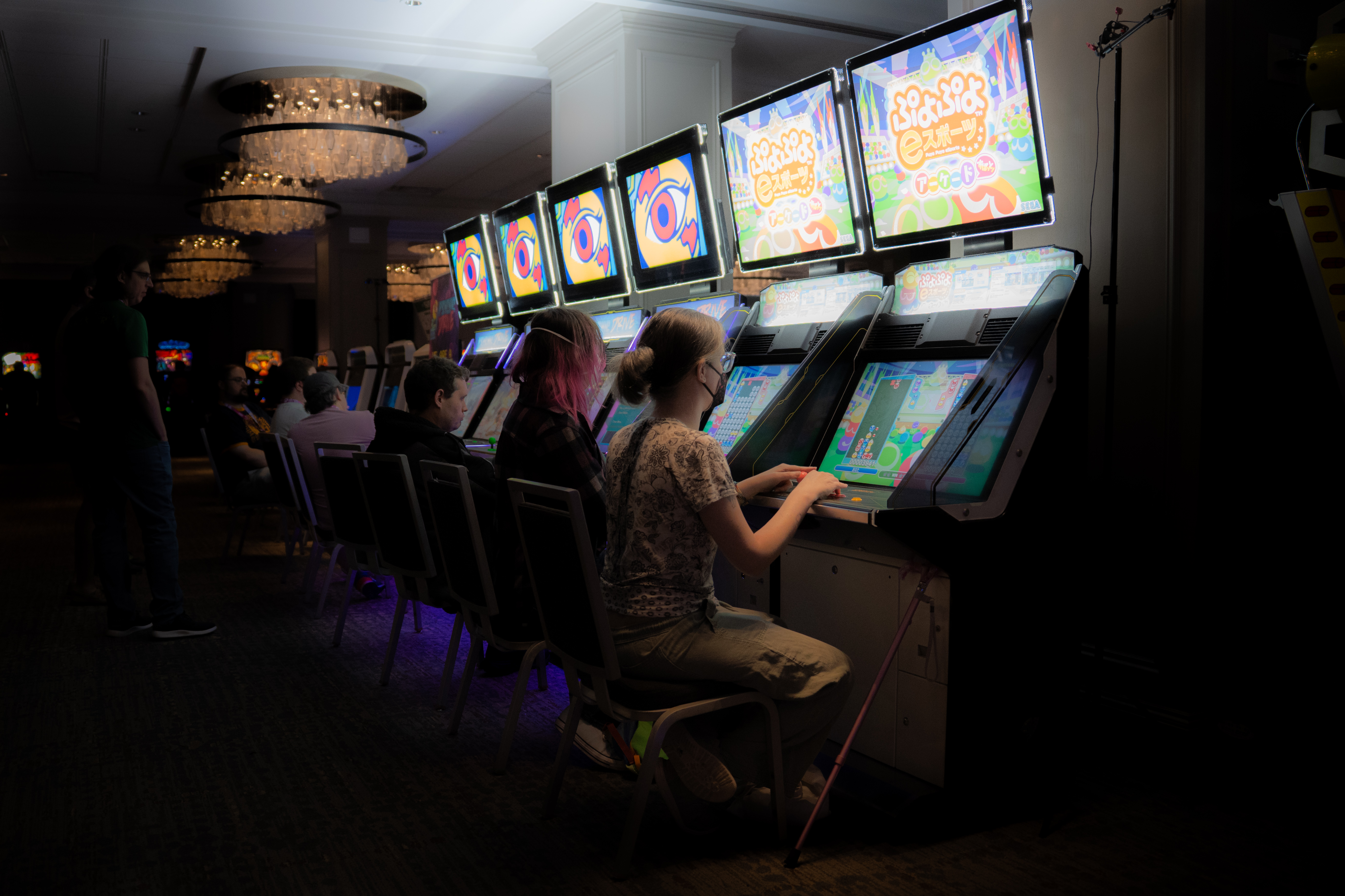 A dark vignette shot of a row of sit down arcade machines, angled so that the machines are disappearing into the distance. sugar sits at the closest machine and is playing Puyo Puyo. To her left is her wife wijn playing the same game of Puyo Puyo on the second machine. They're playing against each other.  - Photo credit: Matt "Lowex" Lowe (official GDQ photographer)