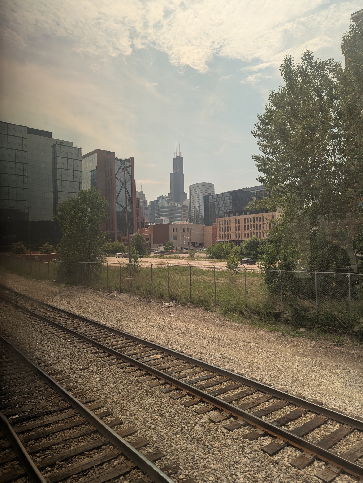 A view of the Sears Tower in Chicago as seen from an Amtrak train window. The window is tinted yellow which gives the whole thing a hazy look. At this point, the smoke had not set in, so this is entirely the window tint making it look that way. In the foreground you can see two train tracks that are used by Metra trains. Beyond that is a small field, a road, and then buildings, with the Sears Tower looming above everything in the background.