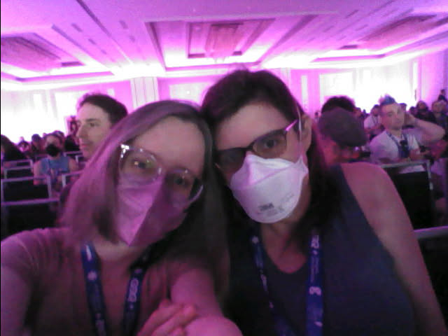 Low res selfie of me and my wife leaning against each other in the audience.  Behind us are more people and a bright pink light at the back of the large ballroom.  I am in my pink KN95 face mask and she in her white N95.
