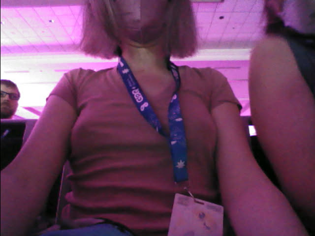 Low res selfie taken from the point of view as if you were in my lap, looking up at me. My boobs look really nice, and you can get a view of the lanyard around my neck. Over my right shoulder, the left side of the photo, a man's head leans into frame looking disappointed or disapproving.