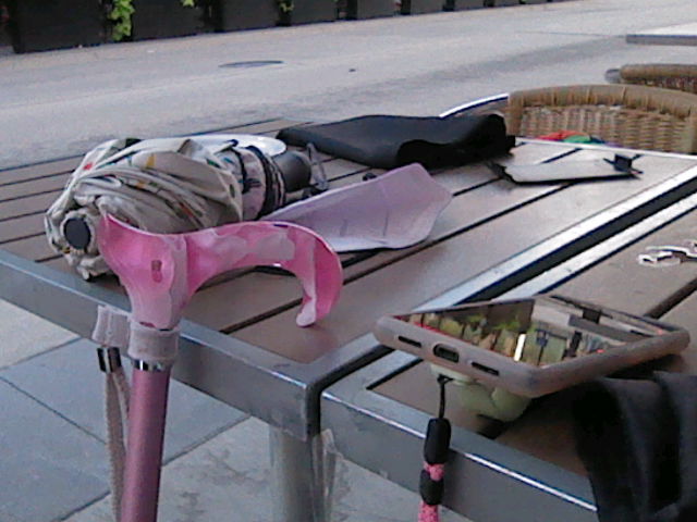 Photograph of my pink cane hanging on an outdoor dining table at a restaurant. Next to the cane are my phone and my parasol. The pink acrylic handle of the cane is a deep pink with white swirls in it. The body of the cane is also pink.