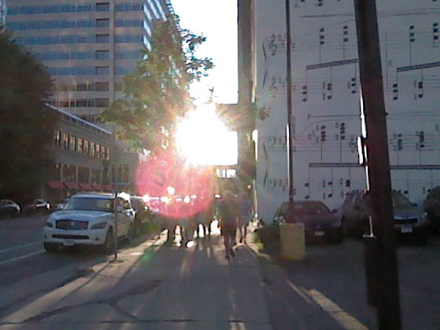photograph of a sidewalk facing into the sunset.  there is a large bloom on the sun and lens flare effect.  to the right is a building with sheet music painted on the side