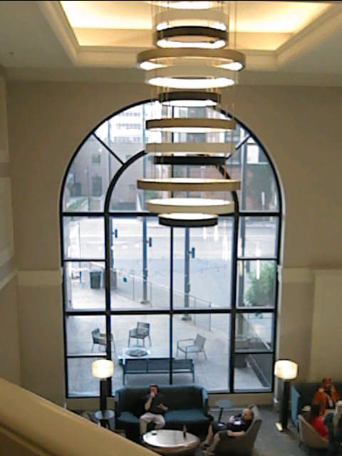 photo graph of a large hanging chandelier in the lobby of the hotel, made up of rings that hang below each other. behind it is a large window with a half-circle shape at the top.