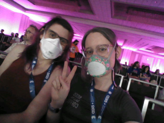 Selfie of my wife and I in the stream room again. I am wearing a flo mask with cherry blossom art on the rim. We are leaning into each other and I am flashing a peace sign with my fingers.