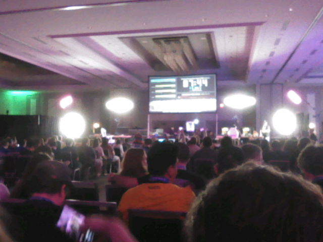 Blurry low res photograph of the main stage. There are a lot of stage lights that are just blown out. You can see the main projection screen with the countdown timer on it, but it is not in focus. You can also see the backs of the heads of audience members.