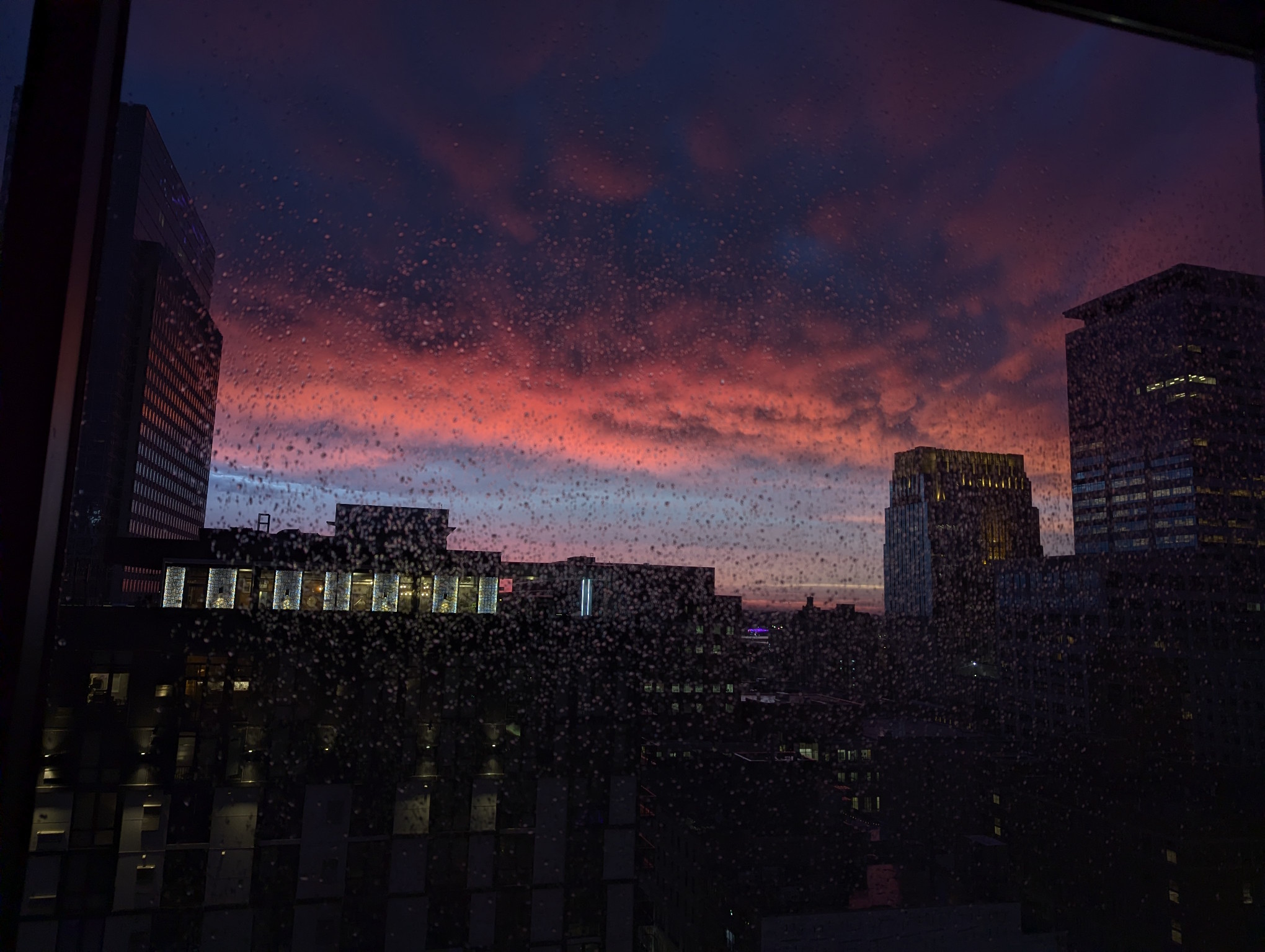 Dark nighttime photograph of an after-thunderstorm sunset over some buildings in downtown Minneapolis. The sky in the distance is blue, and as you look closer there's a shelf of clouds. The bottom edge of the shelf is a bright red that fades into a dark purple almost. The buildings are very dark.