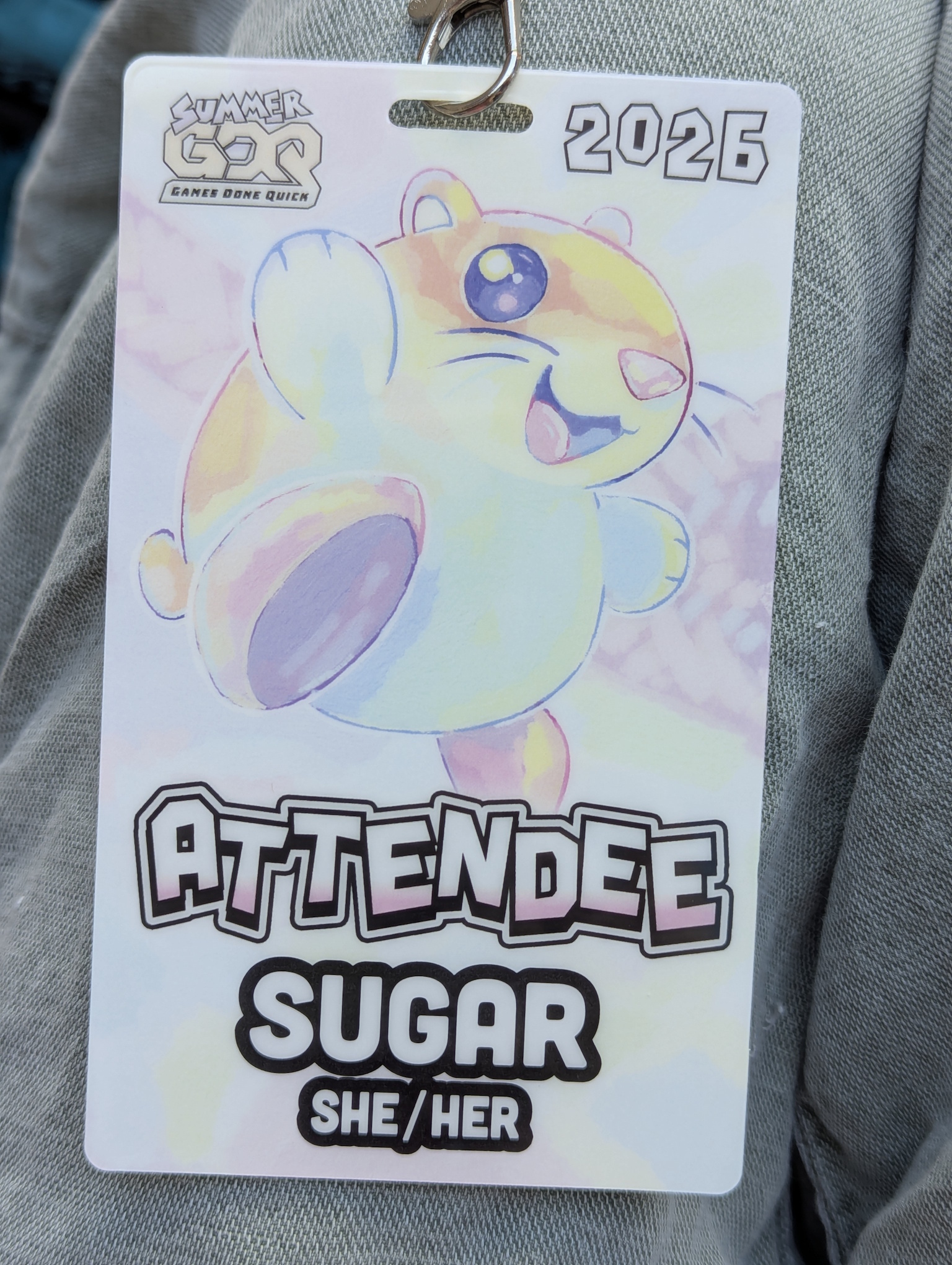 A photo of my SGDQ 2026 badge.  It says ATTENDEE, sugar, and she/her.  The art this time is a light pastel drawing of Rick the hamster from Kirby's Dreamland 2 for the Gameboy.  He is wearing some kind of shoes instead of having actual hamster feet.  Otherwise he's like jumping and looks so happy!