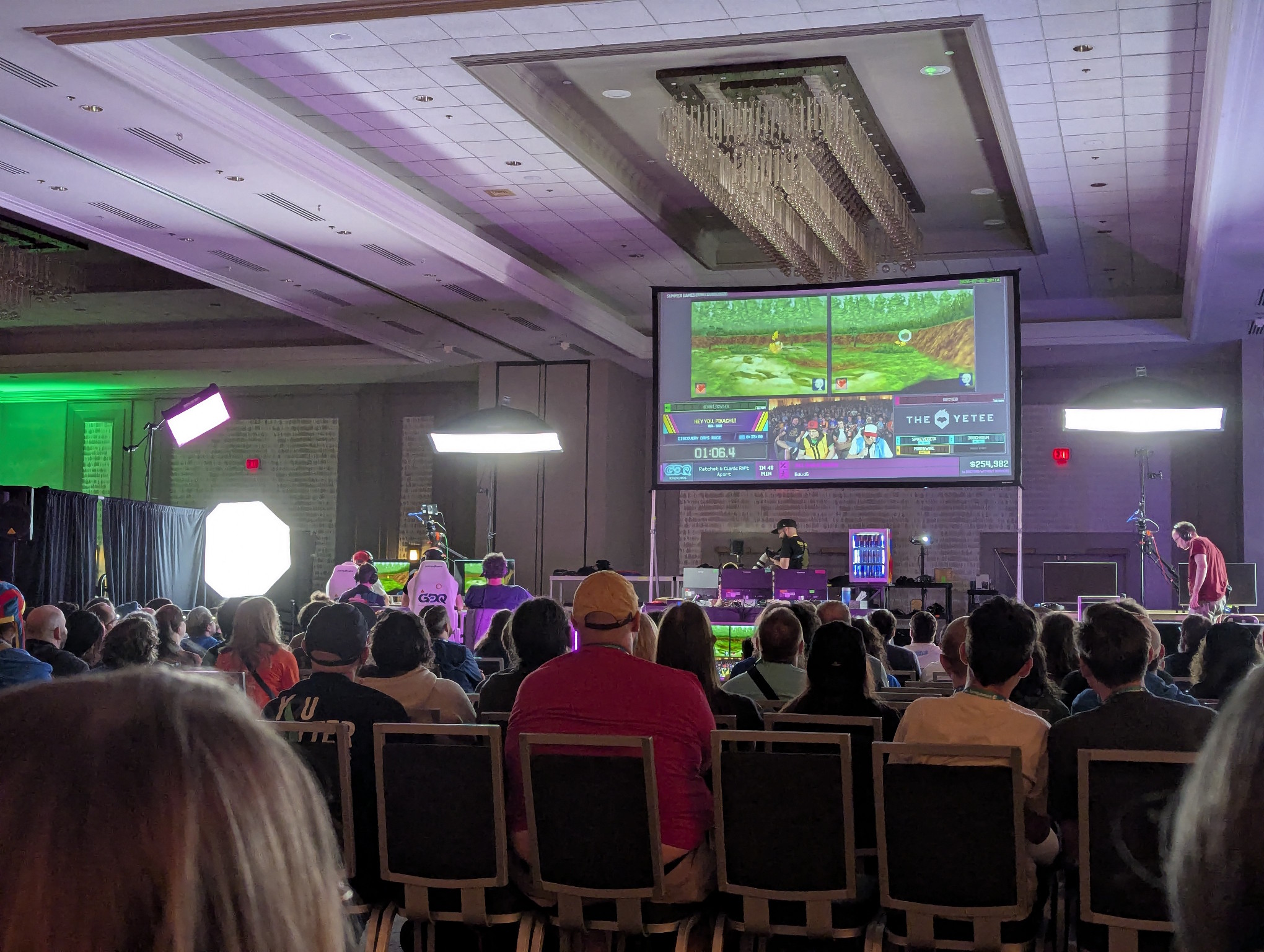 A very clear photograph of the main stage in the stream room. The lights are no blown out and you can see the main projector screen. On it is a two player race of Hey You Pikachu! for the N64. Both players are in a green field trying to talk their Pikachu into doing a specific activity. You can see the audience in front of us.