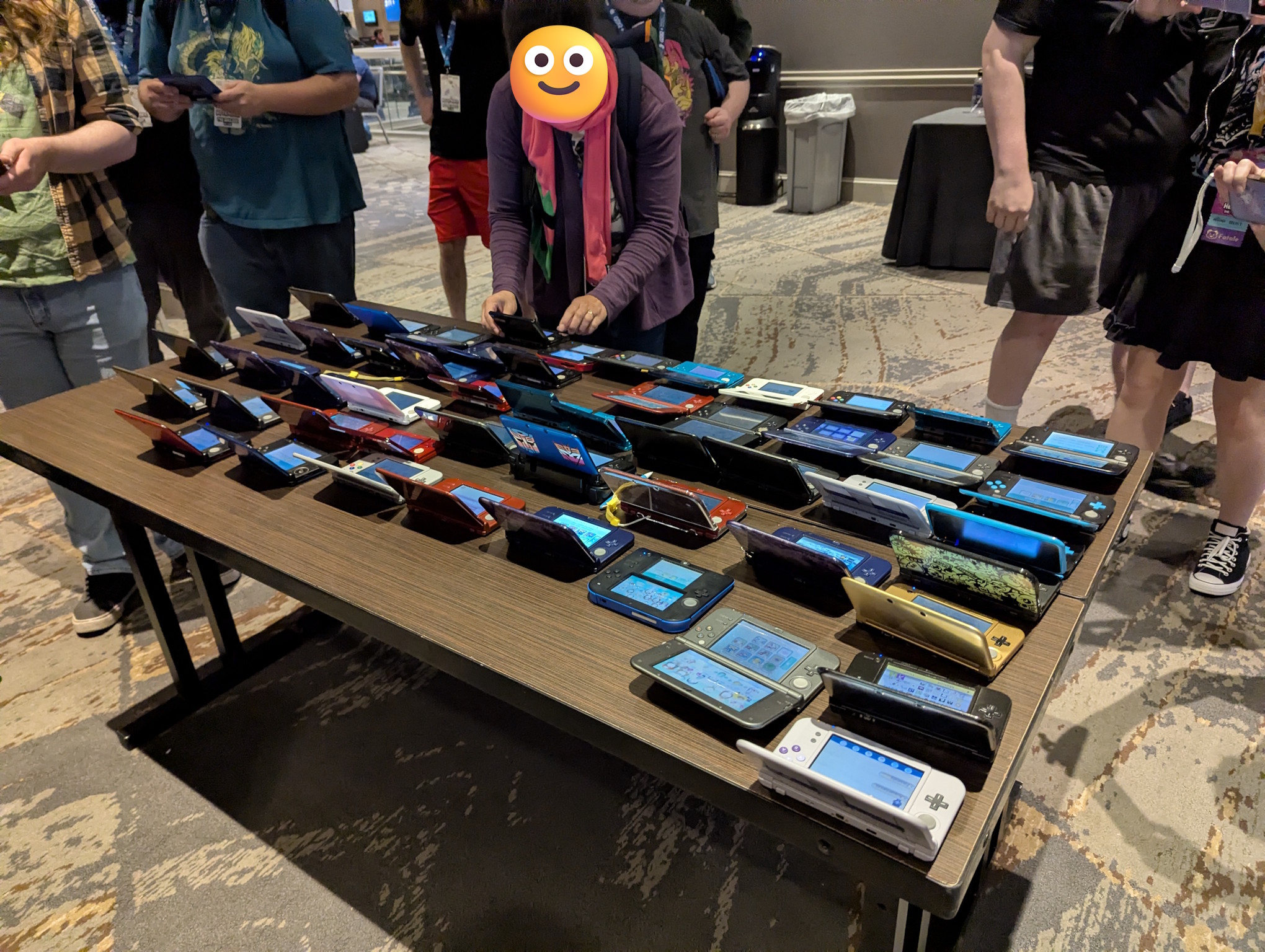 A photograph taken from behind and slightly above of a table full of various Nintendo 3DS systems, set down in order to be professionally photographed.  Around them are a bunch of people looking on and some also taking photos.
