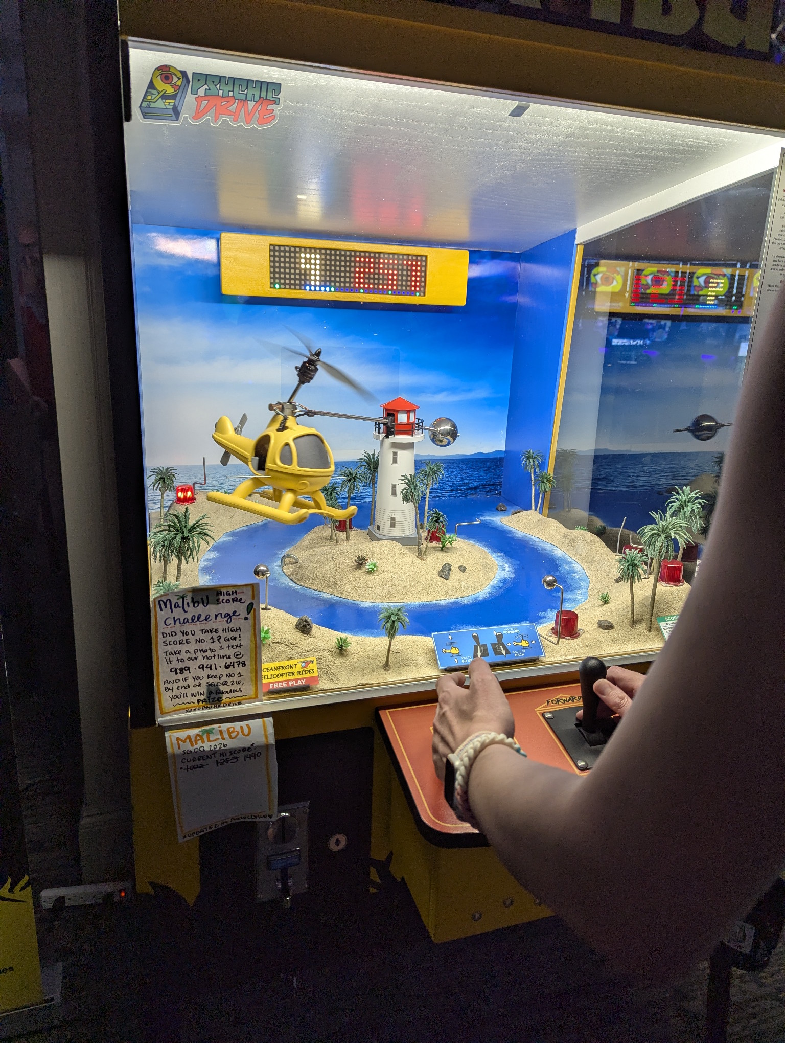 My wife playing a weird small indie arcade game built entirely of physical components. There is a yellow helicopter on an arm that flies around a lighthouse. The scene is tropical, with palm trees and sand and bright blue water.