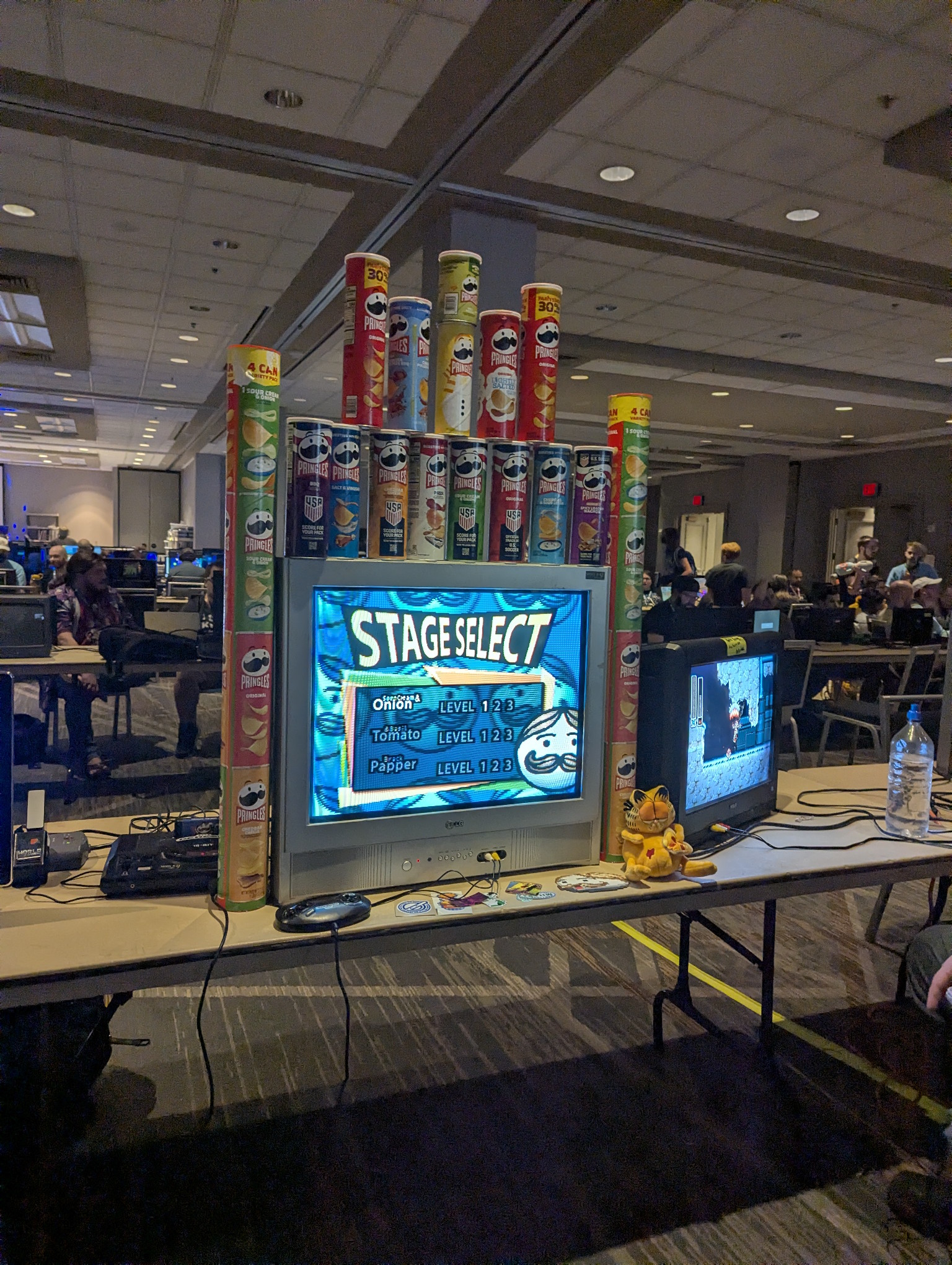 A CRT on a folding card table showing the Stage Select screen of some kind of Pringles branded video game.  Stacked on top of and around the tv are pringle cans.