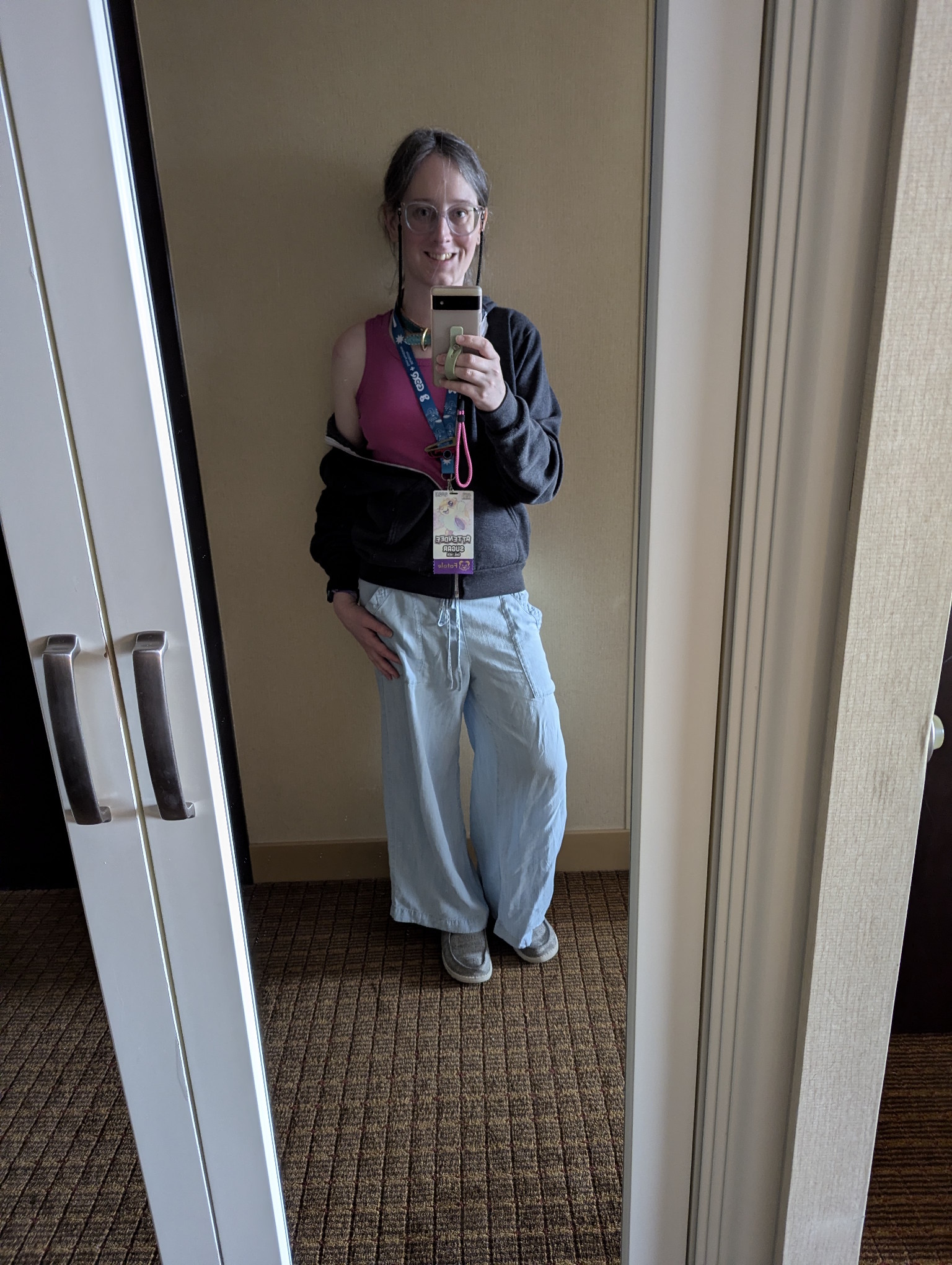 Mirror selfie in our hotel room.  I'm wearing light blue linen pants, a deep pink tank top, and a dark grey hoodie.   I'm smiling.  I have a black glasses chain dangling from my glasses that goes behind my neck.  I'm wearing my badge.