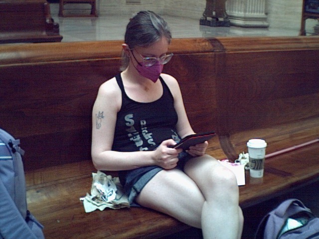 Low resolution photograph of a white woman sitting with one leg over the other on a wooden pew in the waiting area of Chicago's Union Station. She is wearing a pink KN95 face mask and is playing on a red 3DS with coffee and food wrappers next to her. She's wearing a black tank top and jean shorts. Her legs look very muscular somehow!